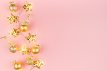 Fototapeta na wymiar Christmas lights and decoration - golden stars garland glowing on soft light pastel pink background with balls, ribbon, border, top view.