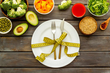 Diet concept. Fork and knife tied with measure tape on plate near vegetables on dark wooden background top view
