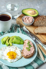 Healthy breakfast with cooked egg, chopped avocado on a plate, cereal bread with pate and coffee with a glass of water