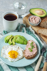 Healthy breakfast with cooked egg, chopped avocado on a plate, cereal bread with pate and herbs and a glass of water with coffee