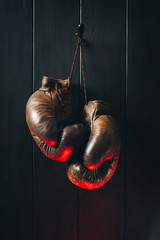 Old brown boxing gloves, hanging on black wooden wall in red lighting. 