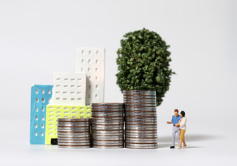 A miniature couple standing with a pile of coins in front of a miniature building.