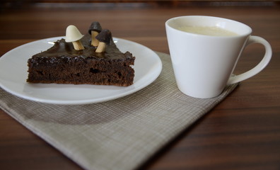 Morning breakfast. On the table is a white ceramic cup with late, on a plate is a chocolate brownie dessert with mushrooms.