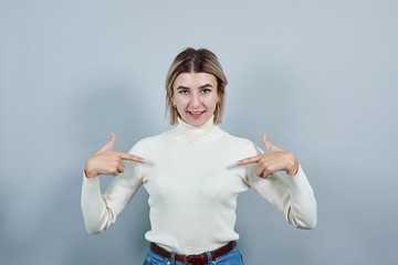 Portrait of smiling pleasant young woman in pastel sweater showing fingers on chest, heart isolated...
