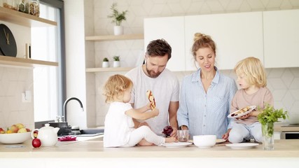 Young family with two small children indoors in kitchen, eating pancakes.