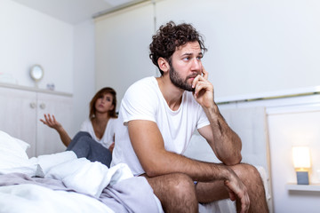 Beautiful girl and a frustrated man sitting in bed and not looking at each other. Upset couple ignoring each other. Worried man in tension at bed. Young couple angry with each other after a fight. - obrazy, fototapety, plakaty