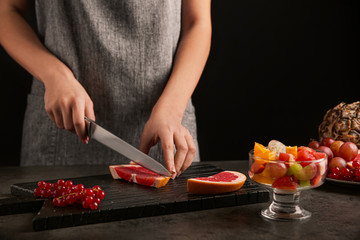 Woman preparing tasty fruit salad on dark background