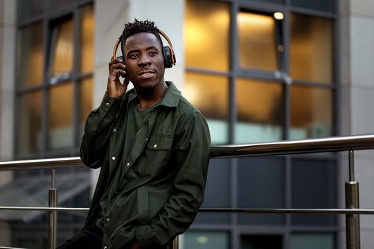 african-american man in stylish jacket in wireless headphones listening music on the street of the evening city. space for text