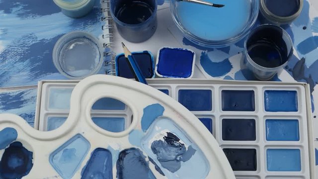 A set of watercolor paints and jars of gouache in the different shades of blue color, the artist palette, a pencil and a paintbrush are laid out on a white surface, still life, background