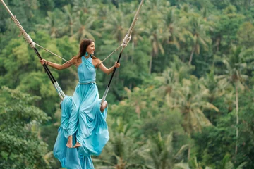 Crédence de cuisine en verre imprimé Bali Belle femme bronzée dans une longue robe turquoise avec un train, chevauchant une balançoire. Au fond, une forêt tropicale et des palmiers. Espace de copie
