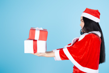 Happy Asian woman in Santa Claus costume holding Christmas gift box on blue background.