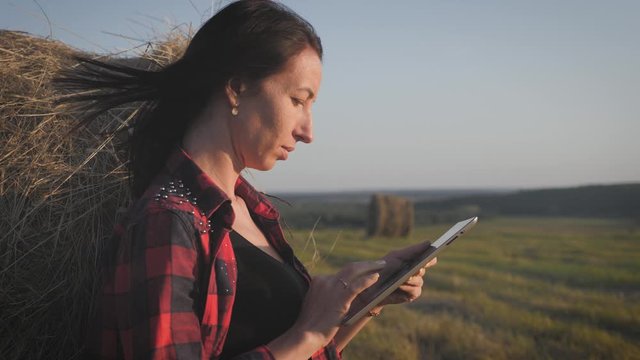 Girl farmer uses tablet in the field next to haystack at sunset. Smart farming, using modern technologies in agriculture.