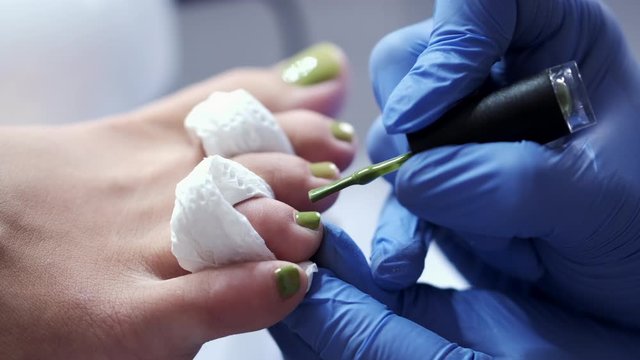 The nails on toes are being polished with a green gel lacquer. The pedicure master is pergorming pedicure in rubber medical gloves.