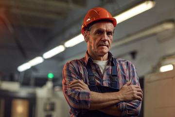 Portrait of confident senior foreman in protective work wear standing with arms crossed and looking...