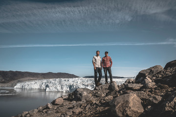 Two young happy male traveler standing in front of Eqip Sermia Eqi glacier in Greenland....