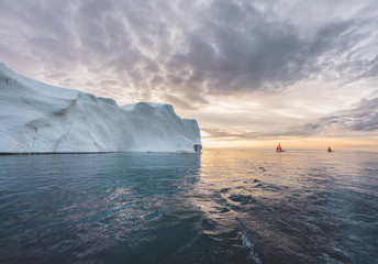 Obraz na płótnie Canvas Beautiful red sailboat in the arctic next to a massive iceberg showing the scale. Cruising among floating icebergs in Disko Bay glacier during midnight sun season of polar summer Ilulissat, Disko Bay