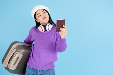 Asian woman wearing sweater carrying luggage shows passport on blue background, travel concept.