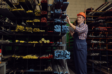 Manual worker in work helmet using digital tablet and checking the metal pipes on the shelves in the factory