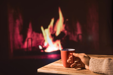 Woman in a winter sweater relaxes sitting near Christmas fireplace with a cup of hot drink and warming woolen blanket. Cozy atmosphere. Winter and Christmas holidays concept