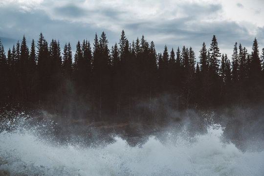 Waves and splashes of mountain river on background of forest, rocks and dramatic sky. Forest river water landscape. Wild river in mountain forrest panorama