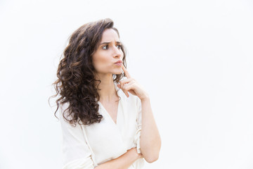 Focused pensive customer touching face with finger, looking away at copy space. Wavy haired young woman in casual shirt standing isolated over white background. Thinking or advertising concept
