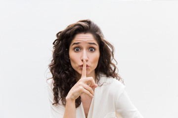 Excited woman with wide eyes making shh gesture. Wavy haired young woman in casual shirt standing isolated over white background. Keeping secret concept