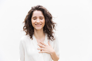 Happy grateful pretty woman with closed eyes applying hand to chest. Wavy haired young woman in casual shirt standing isolated over white background. Relief or gratitude concept - obrazy, fototapety, plakaty