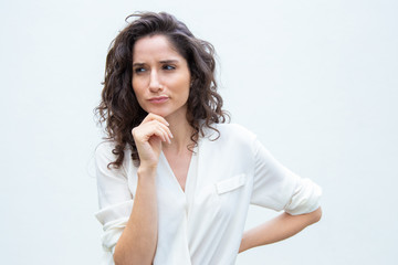 Fototapeta na wymiar Pensive female customer touching chin, looking away, thinking hard. Wavy haired young woman in casual shirt standing isolated over white background. Advertising concept