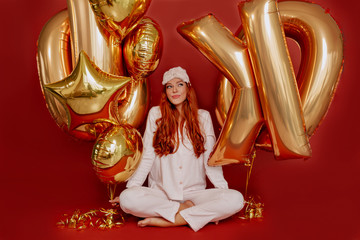 Happy exited lovely red haired woman with long wavy haired wearing pink pajamas posing over red background with gold balloons