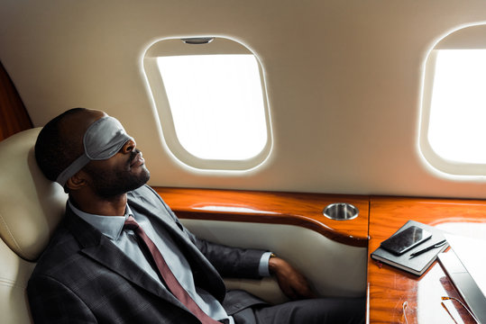 African american businessman with sleeping mask sitting near gadgets in private plane