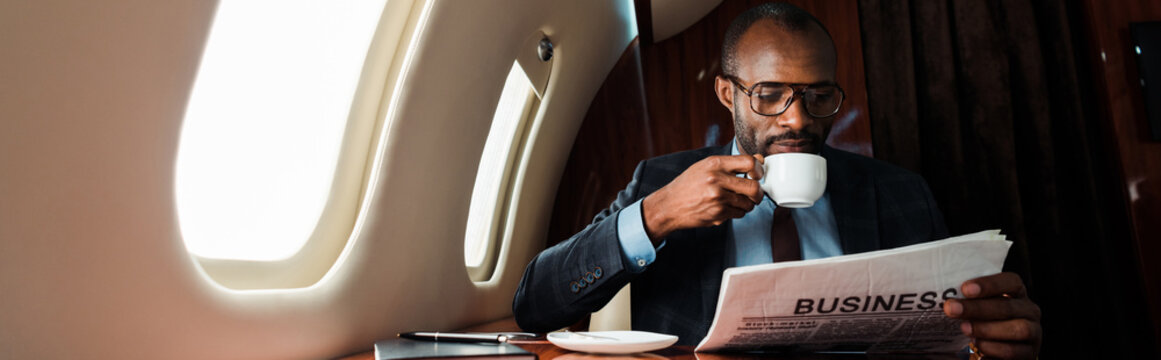 Panoramic shot of African American businessman in glasses reading business newspaper while holding cup in private plane