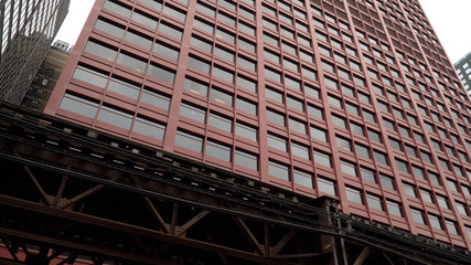 Day time exterior establishing shot of generic large red skyscraper in Chicago. Loop train tracks pass by street level outside building facade for commute to corporate headquarters