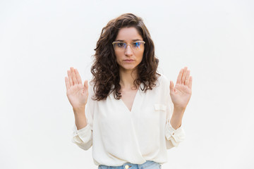 Serious woman in glasses showing palms. Wavy haired young woman in casual shirt standing isolated over white background. Surrender gesture concept