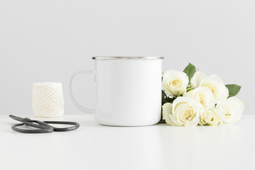 Enamel mug mockup with a bouquet of roses and workspace accessories on a white table.