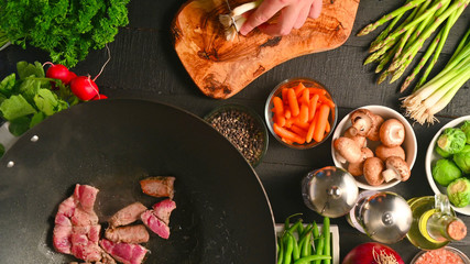 Chef cuts green onions while frying fresh meat in a wok, on a background with vegetables