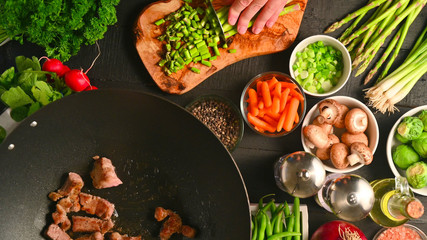 Chef cuts green onions while frying fresh meat in a wok, on a background with vegetables