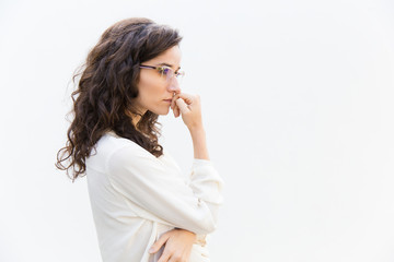 Side of pensive office assistant in glasses leaning face on hand and looking away at copy space. Wavy haired young woman in casual shirt standing isolated over white background. Advertising concept - Powered by Adobe