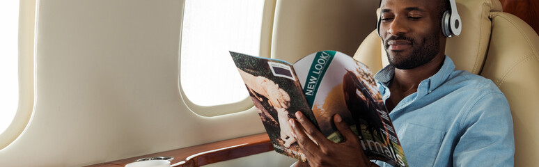panoramic shot of african american man in wireless headphones reading magazine in private plane