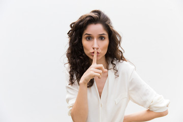 Serious concerned woman showing shh gesture. Wavy haired young woman in casual shirt standing isolated over white background. Keeping silence concept