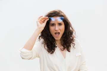 Surprised exaggerated woman taking off glasses, gasping, staring at camera. Wavy haired young woman in casual shirt standing isolated over white background. Conflict concept