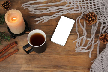 flat lay background with a mug of tea, white smart phone with blank screen, candles, knitted shawl with fringe, coniferous branches and dried orange cinnamon on wood, concept winter mood, copy space