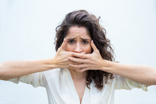Desperate unhappy woman covering mouth with both hands. Wavy haired young woman in casual shirt standing isolated over white background. Keeping mouth shut concept