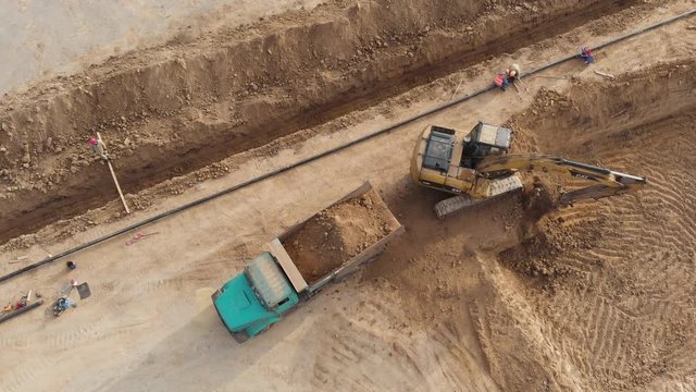 An excavator loads a truck with earth near a dug trench. Aerial view.