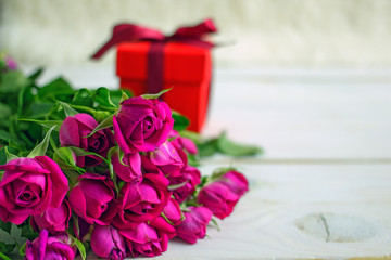 Gift and roses copy space. Blushing gift box with dark red ribbon and bow on a background of red roses on a white wooden table. The concept of  birthday, Valentine's day, mother's day