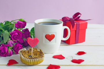 Valentine's Day breakfast. White tea mug with a heart pattern and  homemade cupcake with candles, hearts next to a bouquet of roses and a box with a gift on a wooden table. Copy space