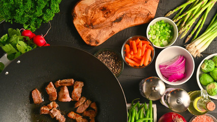 The meat is fried in a wok, against the background of which the cook slices onions. With various vegetables. Culinary background. Flat lay