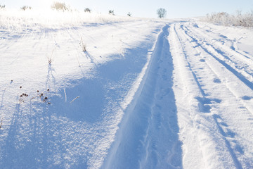 Winter rural landscape with road covered with deep snow and natural sunrays lighting up coverd tall weathered grass.