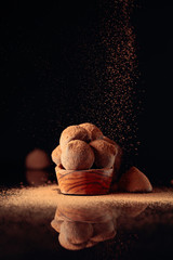 Chocolate truffles in small wooden dish sprinkled with cocoa powder. Black reflective background.