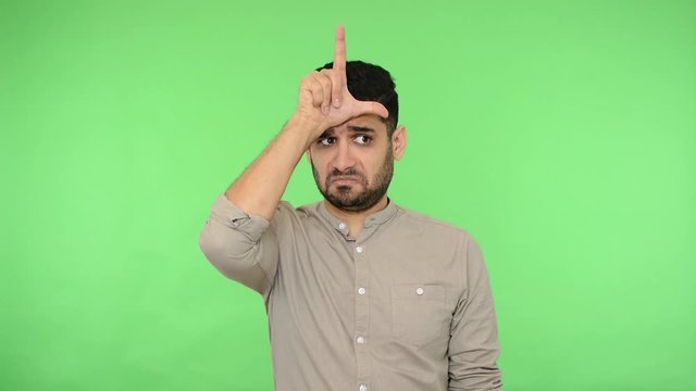 Depressed upset brunette man with bristle in shirt showing loser gesture and looking at camera with dismal expression, feeling despondent about job firing. studio shot, green background, chroma key
