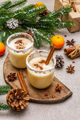 Eggnog with cinnamon and nutmeg for Christmas and winter holidays. Homemade beverage in glasses with spicy rim. Tangerines, candles, gift. Stone concrete background